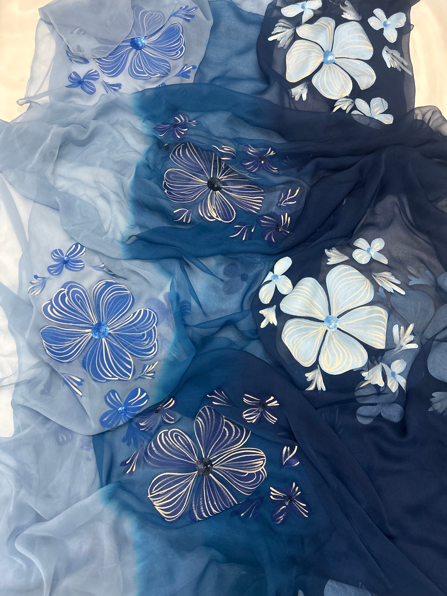 Hand-painted chiffon saree featuring a three-shade blue ombré gradient with large hand-painted light blue flowers and delicate sequin work highlights.